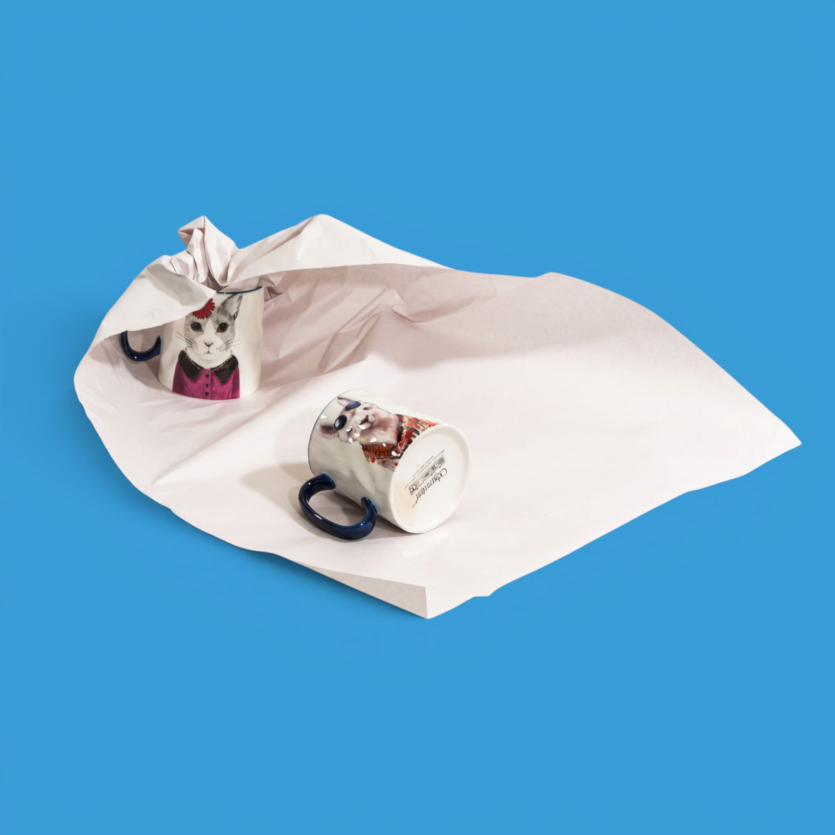 A white ceramic mug with a printed cat illustration and a dark blue handle, shown alongside a crumpled white paper bag on a solid blue background.