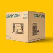 A large cardboard moving box branded ANYVAN, shown at a three-quarter angle on a solid bright yellow background. The box is light brown with teal branding and black line drawings of a window and a front door.