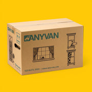 A brown cardboard moving box branded ANYVAN, shown at a three-quarter angle on a solid bright yellow background. The box features green brand lettering, window illustrations, and contact information.